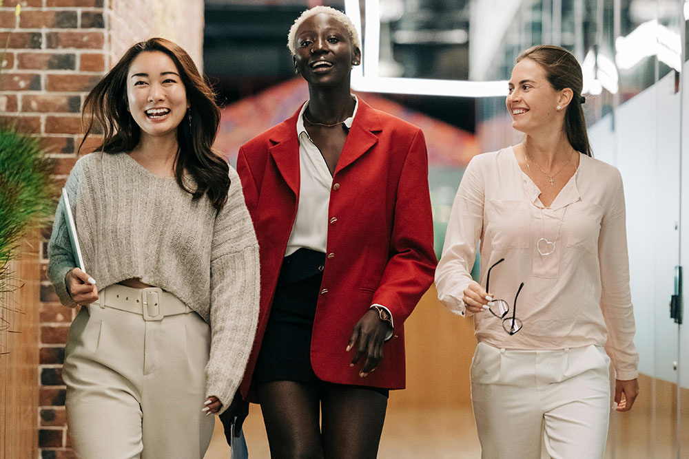 A diverse group of three businesswoman walking confidently and happily down down a corridor together.